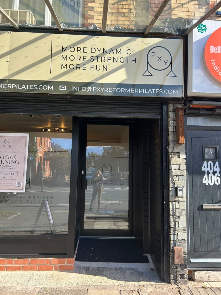 The glass front door of PXY Reformer Pilates in Chorlton, under a sign reading More dynamic, more strength, more fun